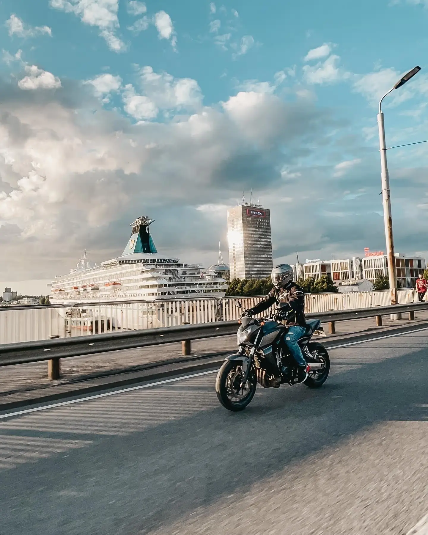 Motociklists ar pilnu ekipējumu gatavojas braucienam pie motocikla, pārbaudot tehniku pirms došanās ceļā.