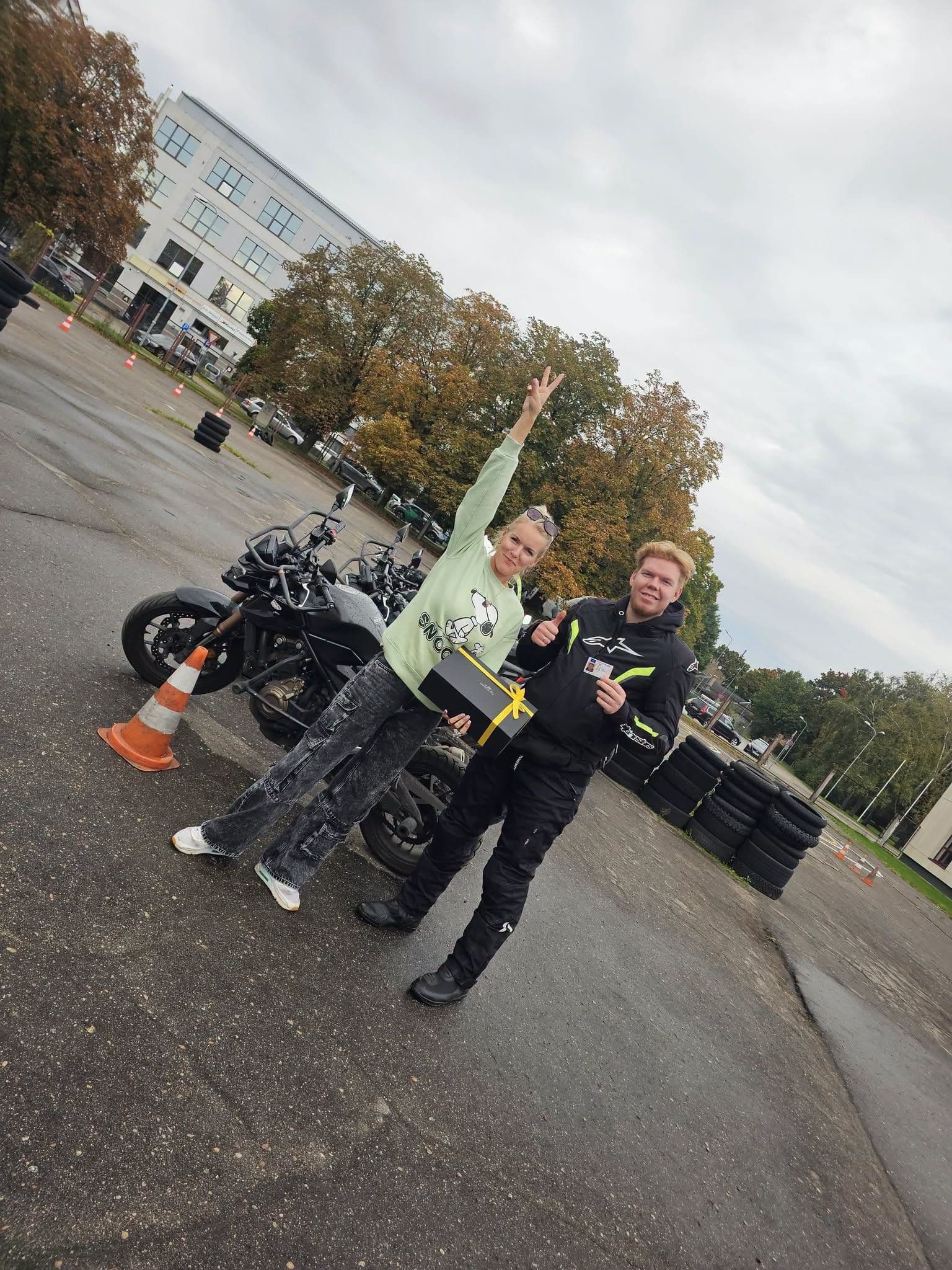 Vīrietis moto ekipējumā smaida, rāda paceltu īkšķi un tur vadītāja apliecību. Blakus sieviete paceļ roku uzvaras žestā un tur dāvanu kasti. Fonā redzami motocikli, konusi un riepu kaudzes braukšanas mācību laukumā.